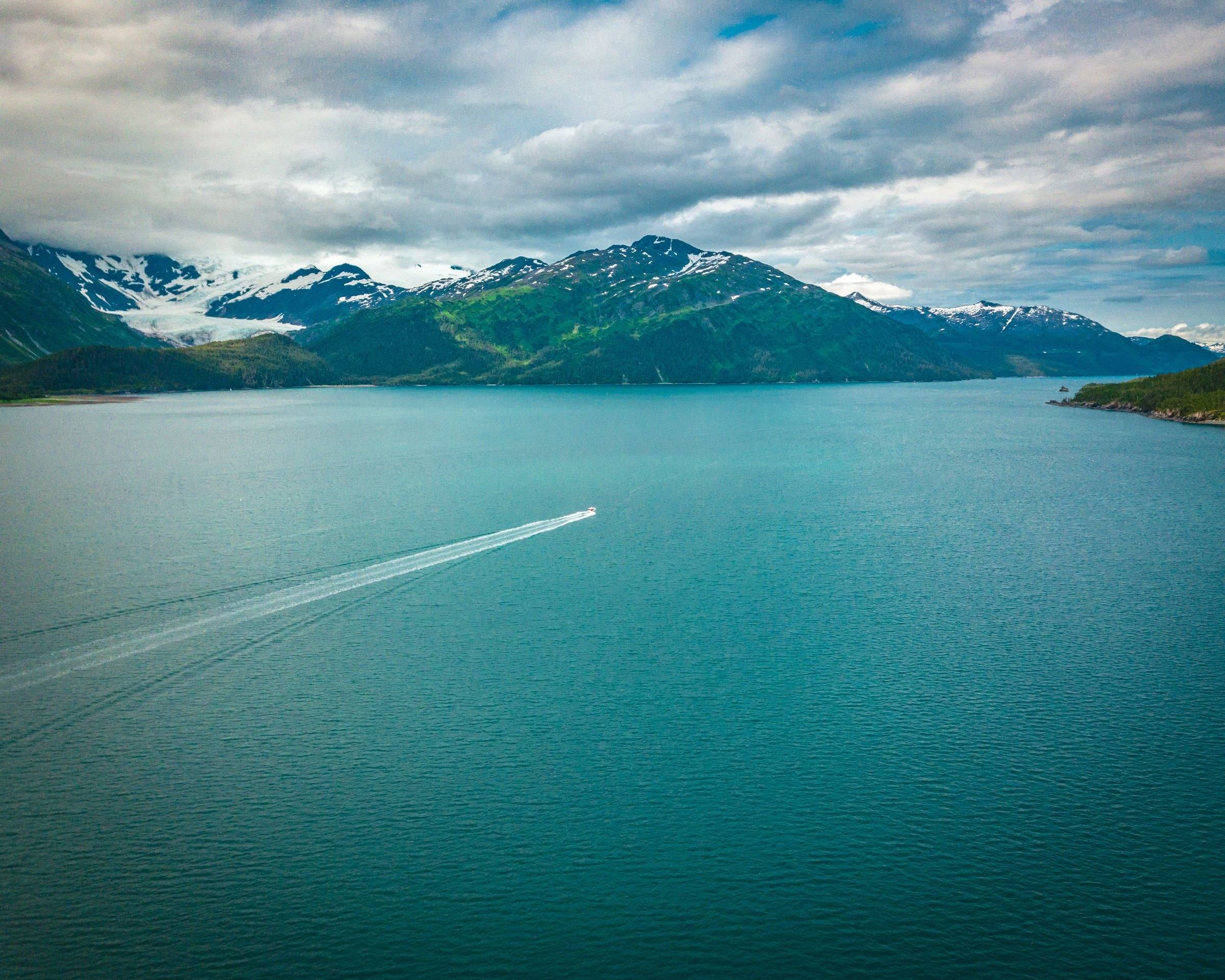 A small boat crossing a teal fjord beneath forested mountains and a glacier